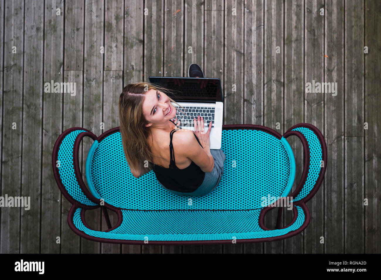 Lächelnde junge Frau sitzt auf Türkis Tisch auf der Terrasse mit Laptop, Ansicht von oben Stockfoto