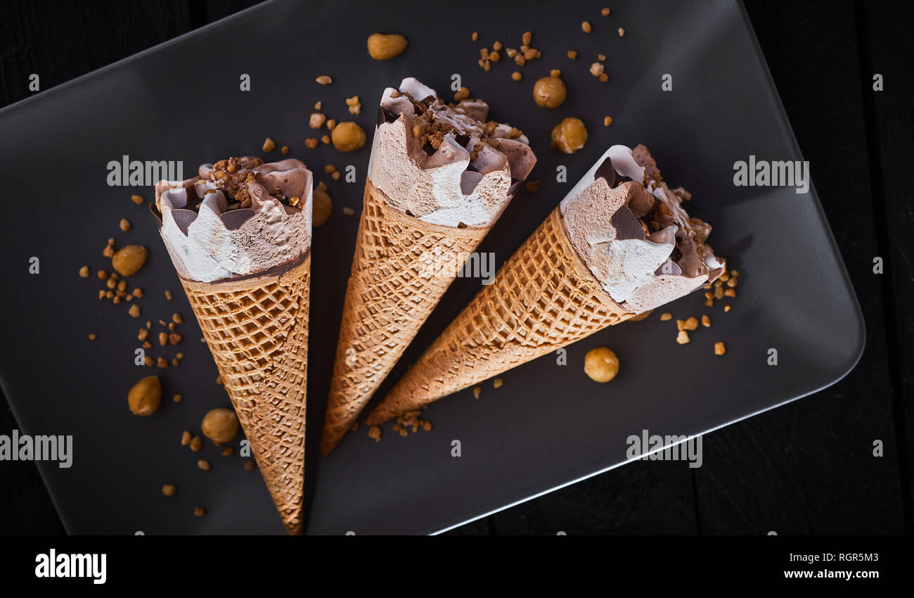 Drei gefrorenes Eis Desserts in Zucker Kegel gekrönt mit Schokolade und mit verstreuten gehackte Nüsse auf einem schwarzen rechteckigen Platte serviert. Stockfoto