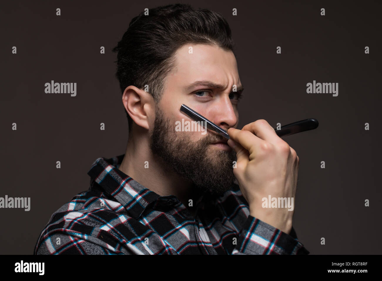 Stattlicher Mann mit langem Bart halten ein Rasiermesser biegen das Gesicht tragen in Plaid Shirt auf grauem Hintergrund Stockfoto