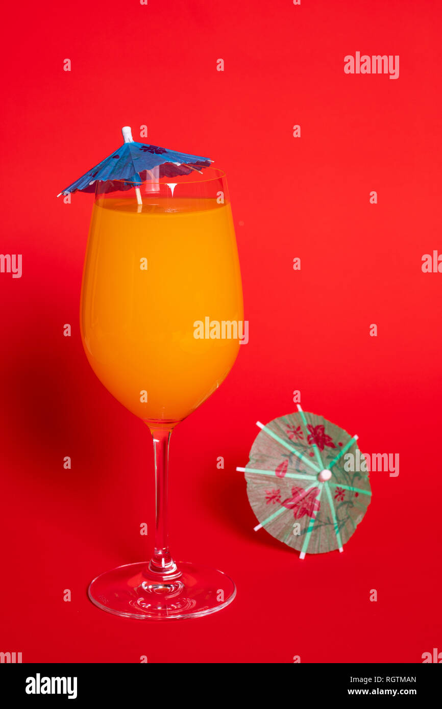Orange Drink mit einem blauen Schirm in einem Glas Wein gegen eine feste rot hinterlegt. Ein grünes Dach liegt neben dem Glas. Stockfoto