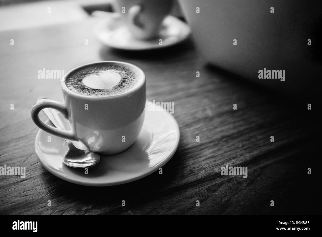 Eine weiße Tasse und Untertasse, silbernen Löffel, mit Kaffee und ein weißes Herz Form latte Schaum Kunst oben gefüllt. Sitzen auf einem hölzernen Tisch, neben einem Laptop. Stockfoto