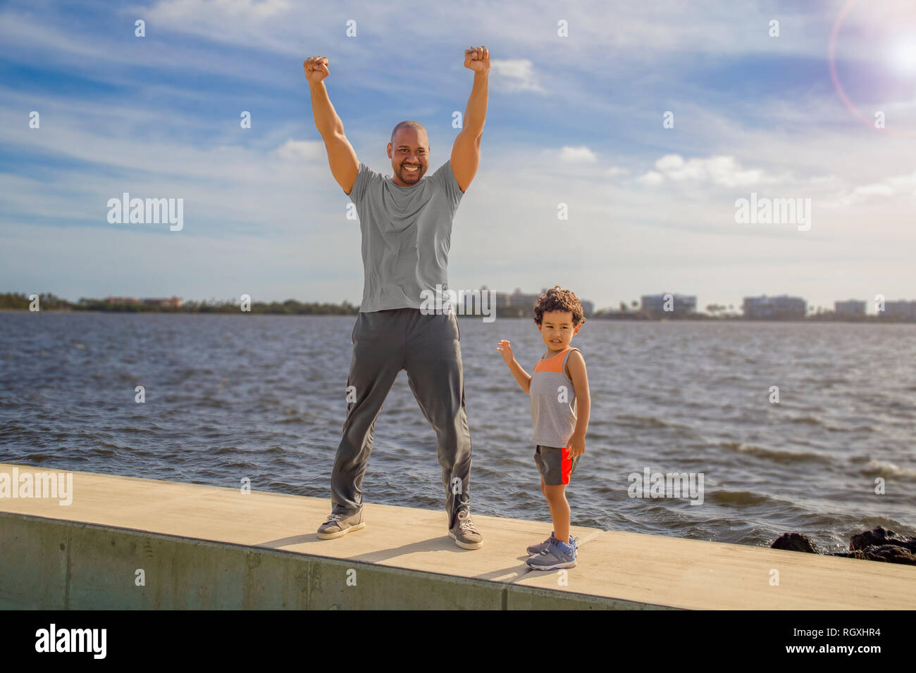 Ein Vater steht hoch über dem Meer an der Wand mit seinem Sohn mit beiden Händen hoch, reckte die kid Sieg für zusammen trainieren in den Morgen zu zeigen. Stockfoto