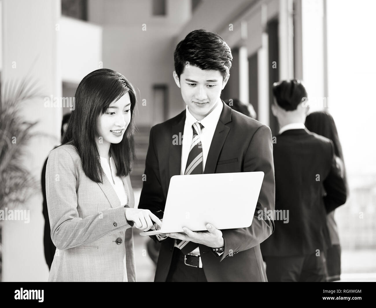 Junge asiatische Geschäftsmann und Geschäftsfrau zusammen arbeiten im Büro mit Laptops, Kollegen sprechen im Hintergrund, Schwarz und Weiß. Stockfoto