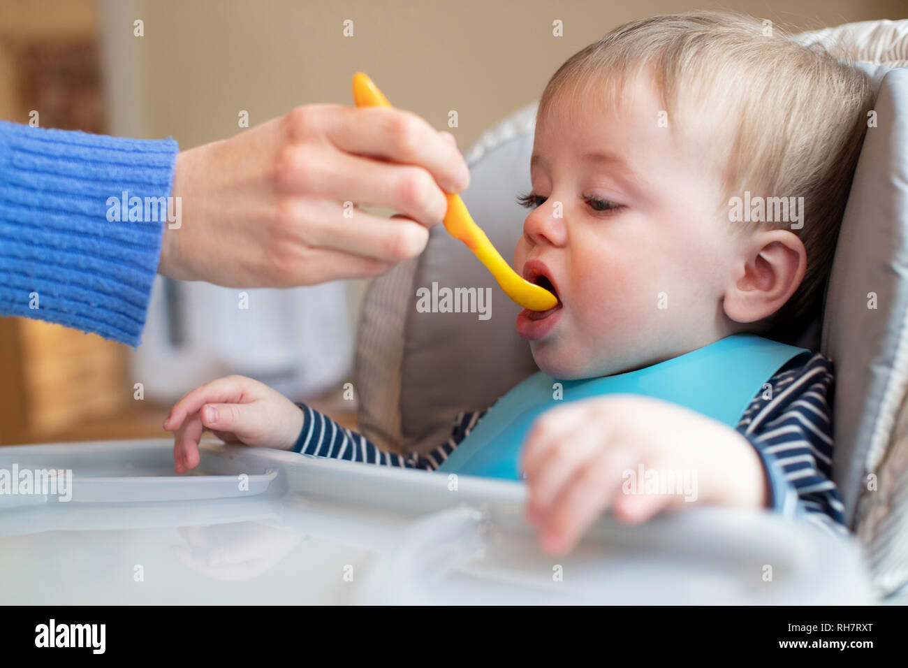 Baby Boy zu Hause in Hochstuhl Zugeführtes feste Nahrung von der Mutter mit Löffel Stockfoto