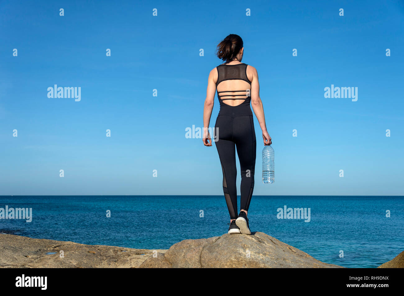 Sportlerin steht auf einem Felsen am Meer mit einem Kunststoff Flasche Wasser Stockfoto