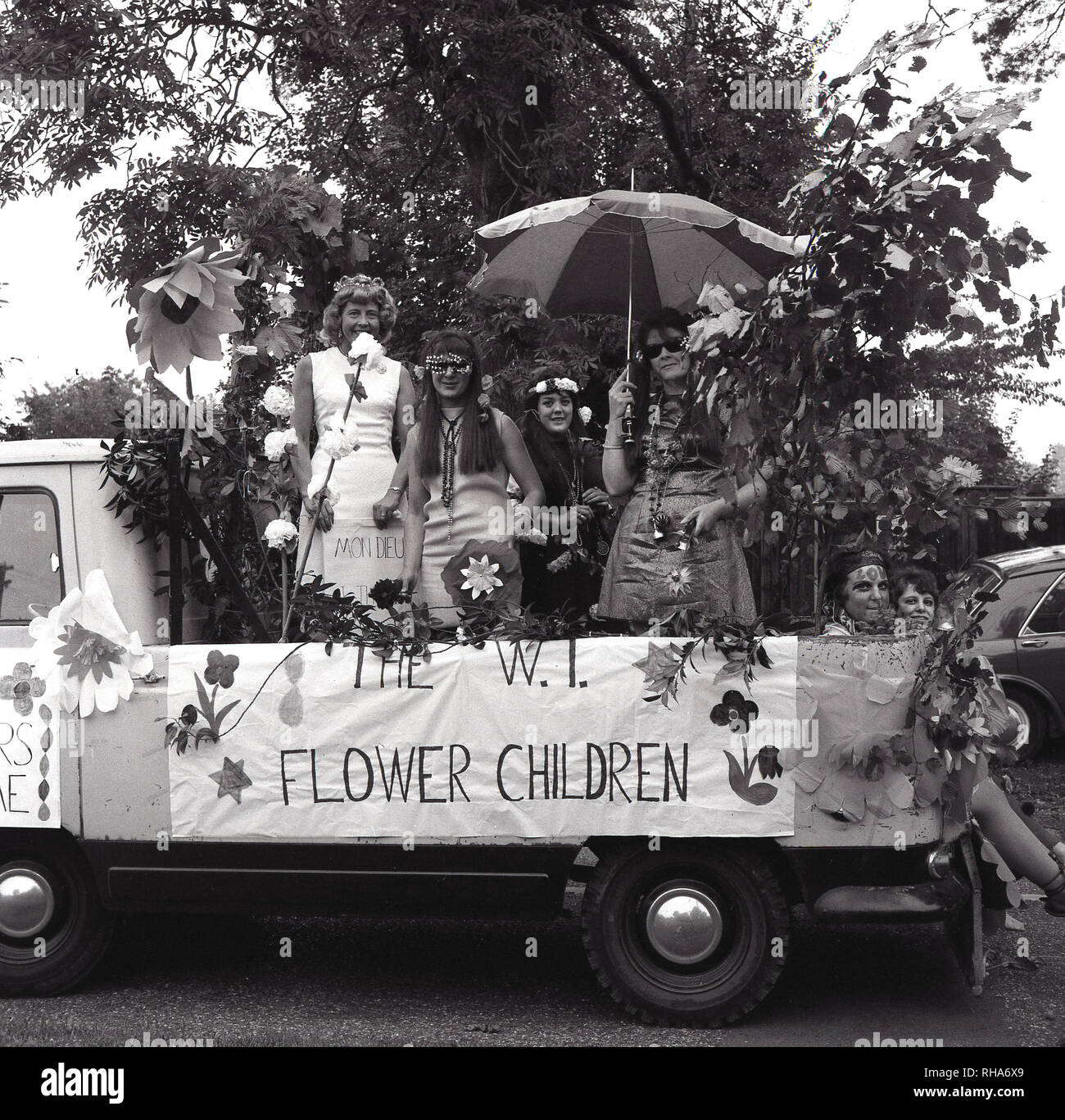 1967, historisch, 60er Jahre 'Flower Power'... Leute, die in einem kleinen offenen Kleinbus oder Pick-up-Truck reisen, einen Wagen, an einem lokalen Karneval oder fest teilnehmen, England, Großbritannien. Der Schwimmer ist mit „Blumen“ verziert und ein Banner an der Seite besagt: Das W.. I Blume Chlldren. Als Symbol für die Konzepte von Frieden und Liebe und unversöhnlicher Zugehörigkeit und verbunden mit der „Hippie“-Bewegung entstand Mitte der 60er Jahre die Blumenkraft als Antikriegsbotschaft. Das Jahr 1967 wird allgemein als „Sommer der Liebe“ bezeichnet. Stockfoto