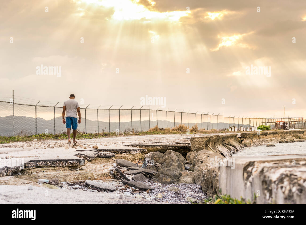 Junge schwarze Mann zu Fuß entlang der Straße am Meer beschädigt, in Richtung Sonne strahlen durch die Wolken. Konzept der Hoffnung, kaputte Straße, Ausdauer Stockfoto