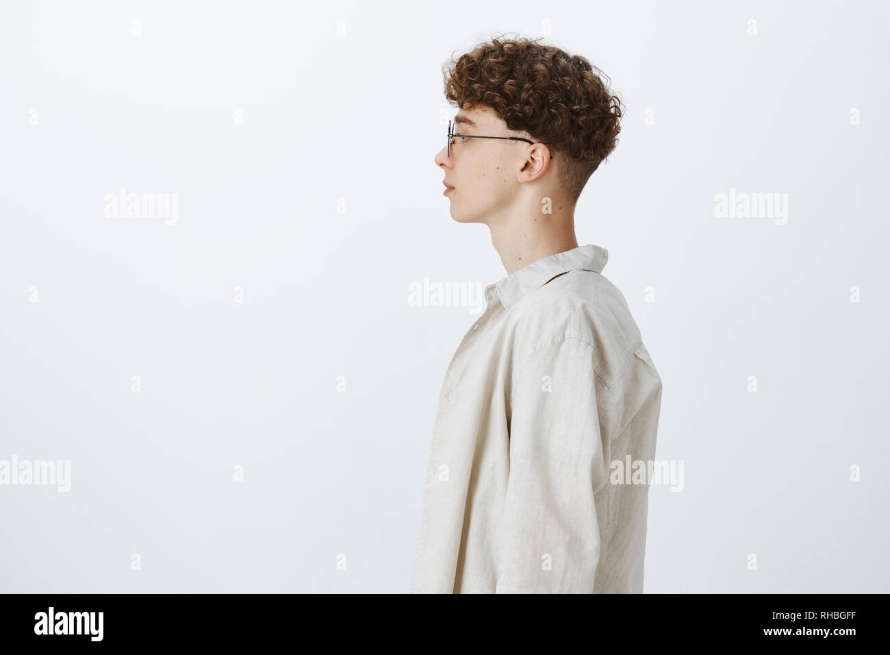 Profil von stilvollen hübscher junger Hipster männliche Freelancer mit Curly Frisur in Gläsern und Links zu schauen und beiläufig stehend über grauer Hintergrund Stockfoto