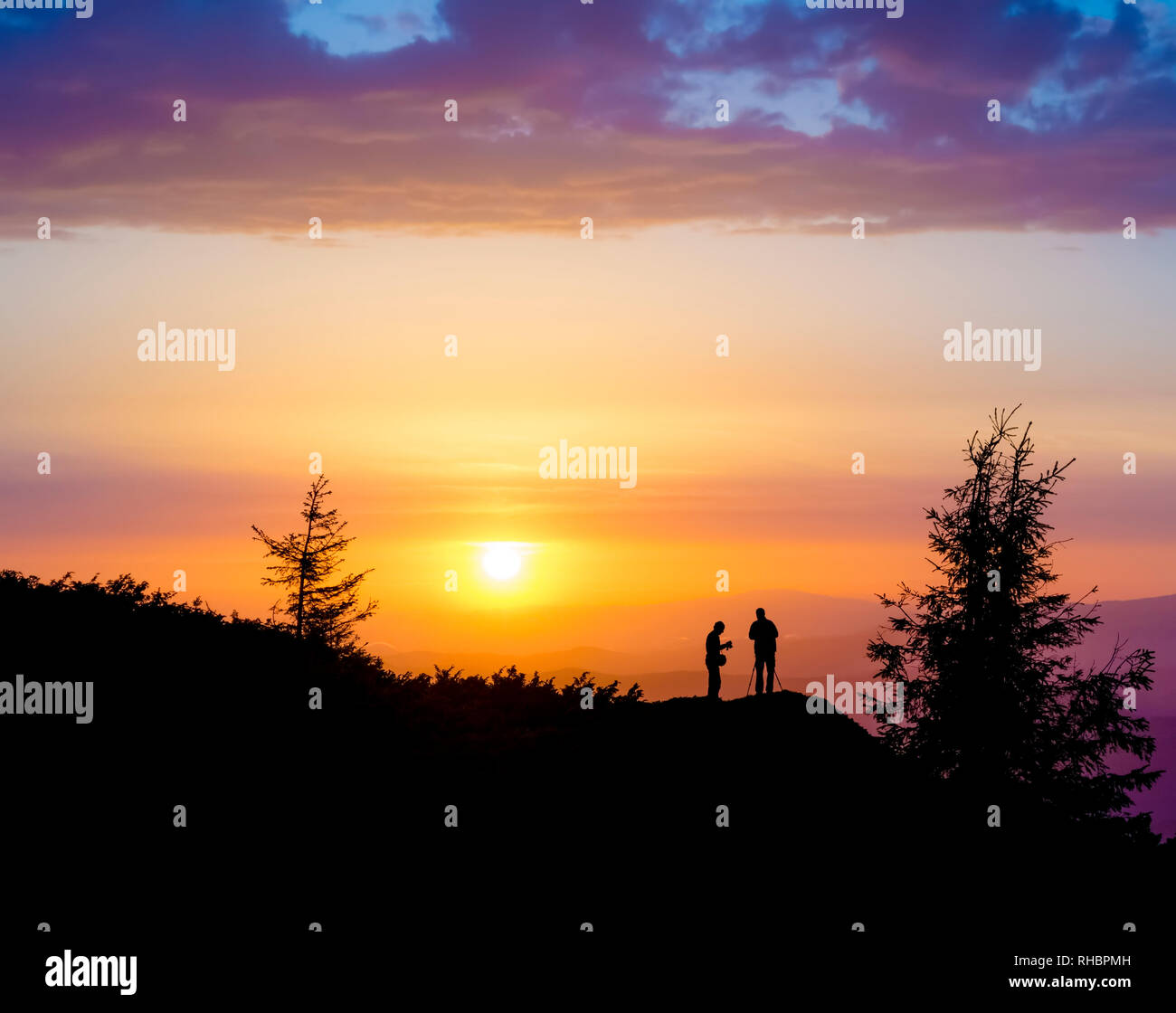 Silhouetten von zwei Menschen, die auf einem Felsen in der Nähe des Baumes und den Blick auf die Sonne. Sonnenuntergang in den Bergen. Sommer in den ukrainischen Karpaten Stockfoto