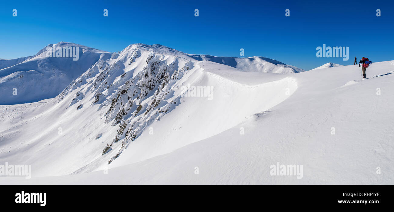 Panorama der schneebedeckten Berggipfel. Scharfe Steine auf einem steilen Hang. Zwei Touristen. Der Himmel ist klar, sonnig. Winter. Die Ukraine Stockfoto