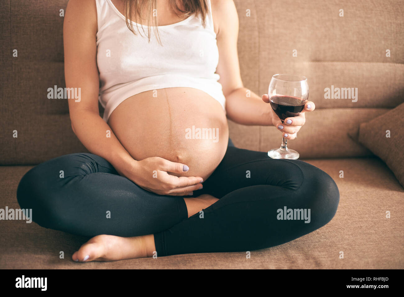 7/8-Ansicht der zukünftigen Mutter in weißem Hemd und schwarzen Hosen sitzen auf einem Sofa zu Hause und trinken Alkohol. Schwangere Frau Wein trinken während der Schwangerschaft. Konzept der Bedrohung, ungesunde Lebensweise. Stockfoto