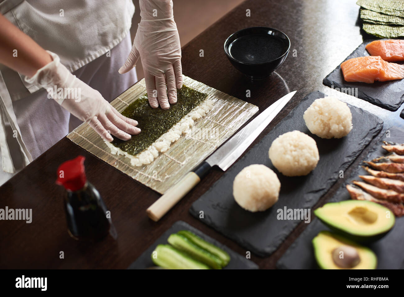 Nahaufnahme der Chef's Hände vorbereiten Rolling Sushi mit Zutaten: Reis, Nori, Avocado, Lachs, Sojasoße Stockfoto
