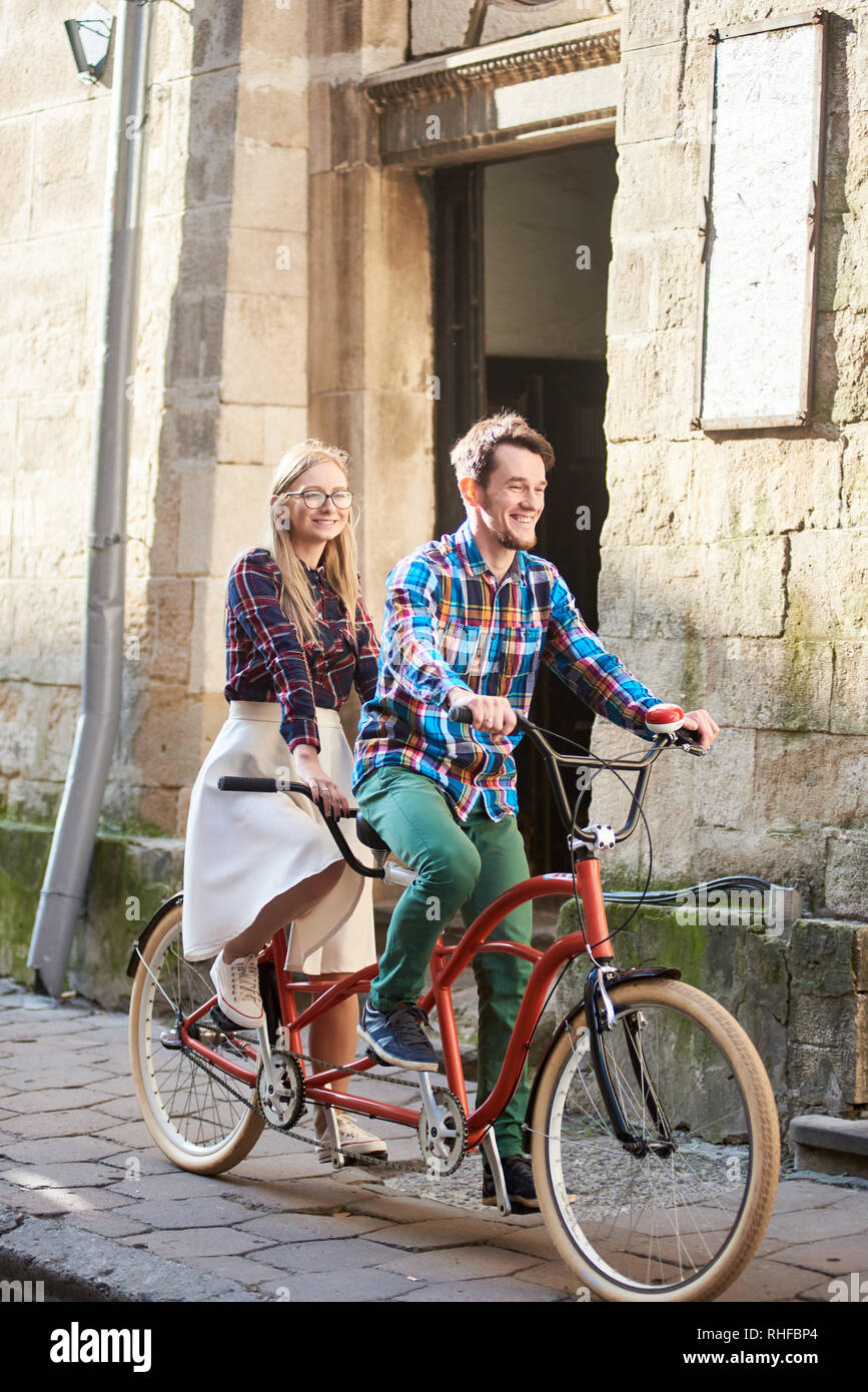 Aktive Ihnen gerne touristische Paar, bärtigen Mann und lange Lächeln - behaartes blonde Frau in weißen Rock und Brille Fahrrad Tandem Bike entlang der befestigten Stadt Bürgersteig auf sonnigen Tag durch alte Gebäude aus Stein. Stockfoto