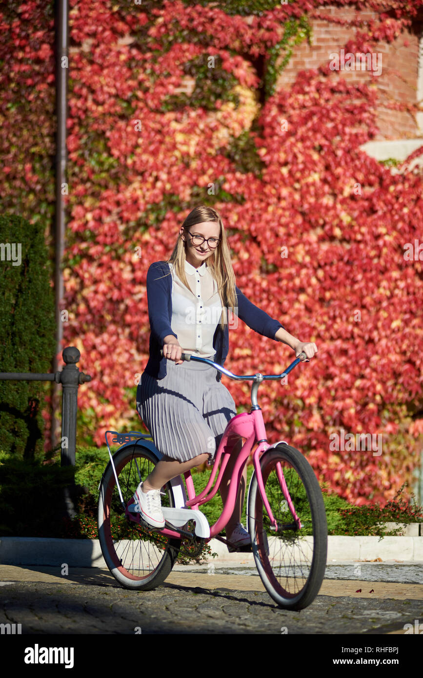 Junge glücklich Langhaarigen lächelnde blonde Frau in Freizeitkleidung und  Gläser Radfahren pink lady Fahrrad auf hellen, warmen sonnigen Tag auf dem  Hintergrund der Ziegelmauer mit schönen roten Efeu Blätter überwuchert  Stockfotografie -