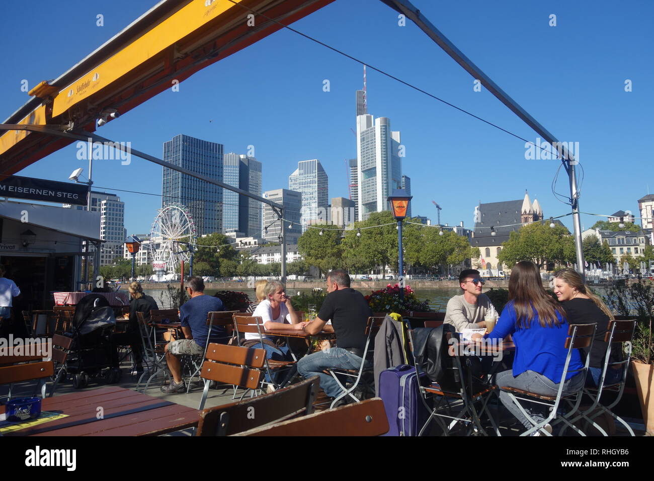 Menschen mit einem Drink auf dem Bootshaus schwimmenden Restaurant und Bar am Ufer des Mains mit der Skyline des Bankenviertels hinter sich. Stockfoto