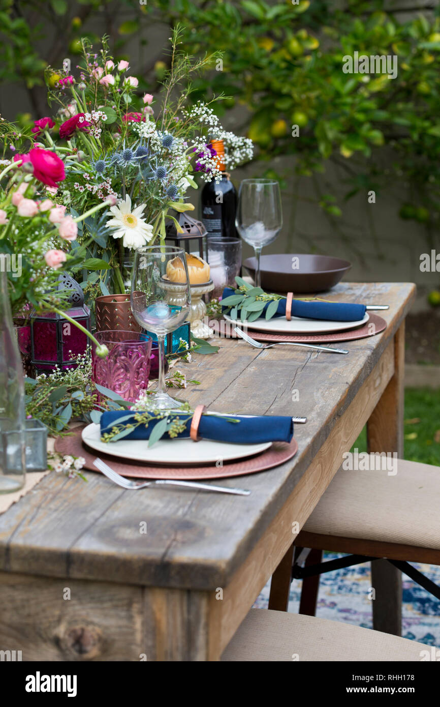 Blau serviette in Ring auf weiße Platte mit Kupfer Ladegerät, setzen Sie die Einstellung auf Holz Outdoor Party Esstisch mit Blumen, Glas, und Kerze. Stockfoto