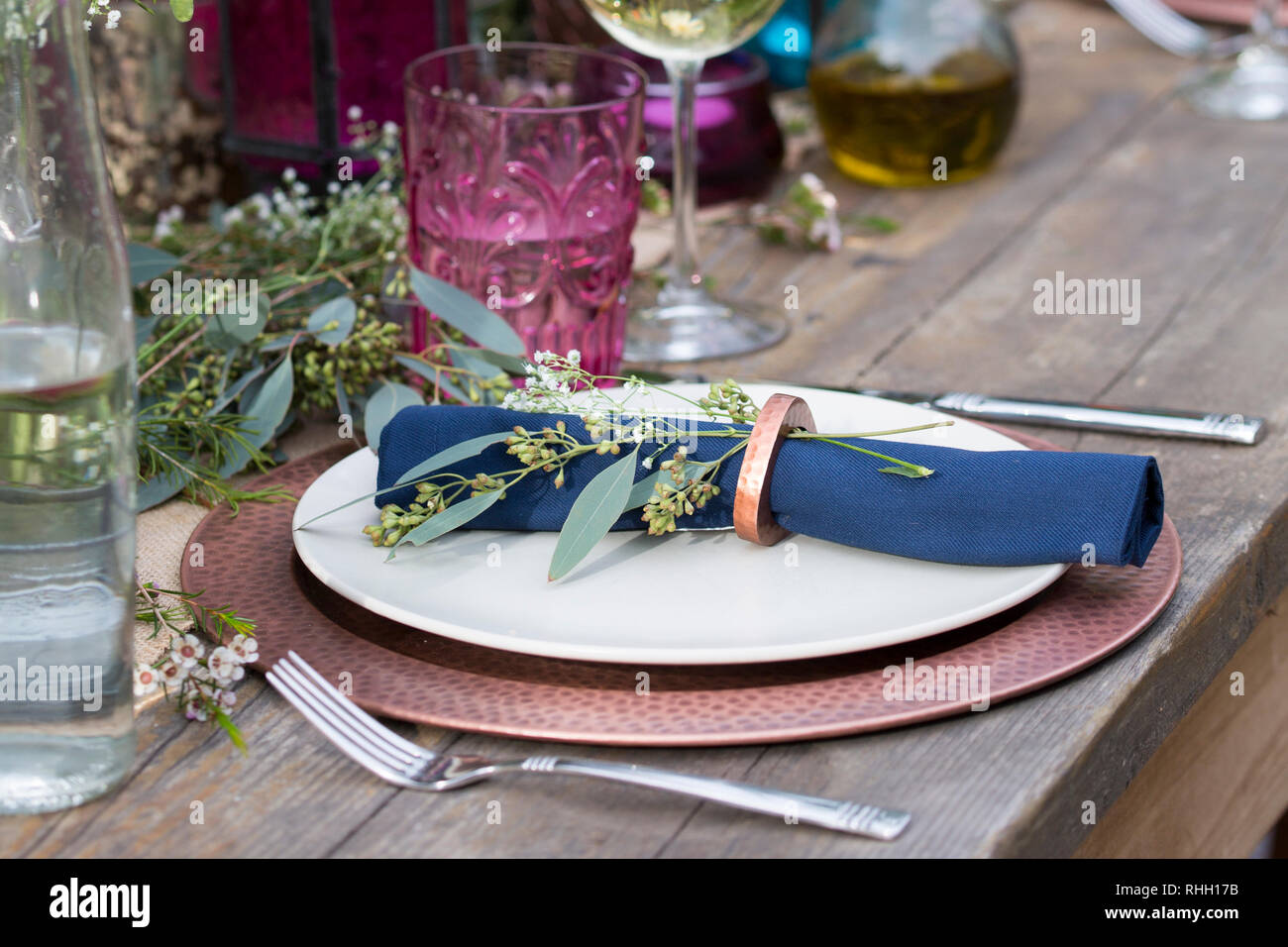 Blau serviette in Ring auf weiße Platte mit Kupfer Ladegerät, setzen Sie die Einstellung auf Holz Outdoor Party Esstisch mit Blumen, Glas, und Kerze. Stockfoto
