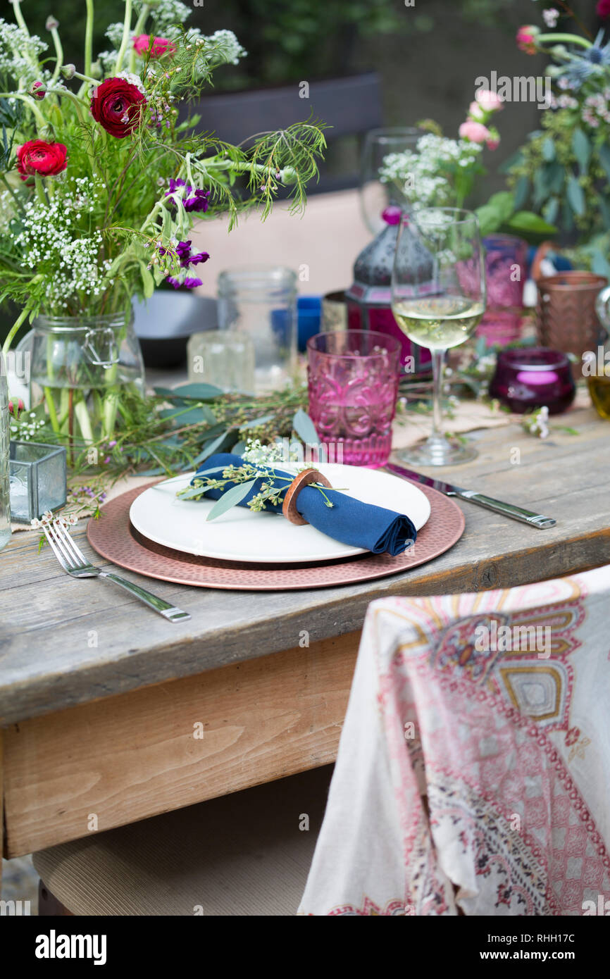 Blau serviette in Ring auf weiße Platte mit Kupfer Ladegerät, setzen Sie die Einstellung auf Holz Outdoor Party Esstisch mit Blumen, Glas, und Kerze. Stockfoto