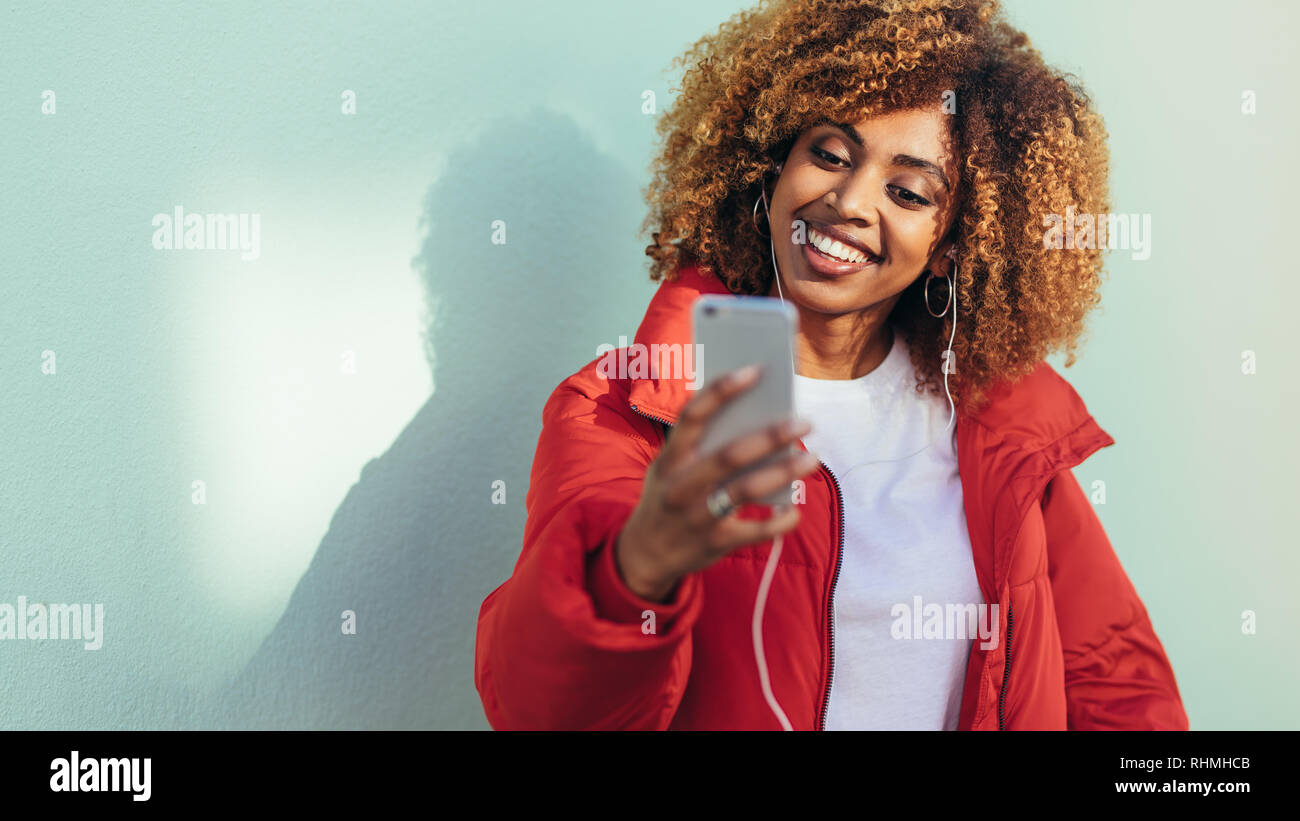 Fröhliche afro-amerikanische Frau an ihr Mobiltelefon suchen, video chat. Lächelnde Frau mit einem selfie mit Mobiltelefon, während Sie Musik hören. Stockfoto