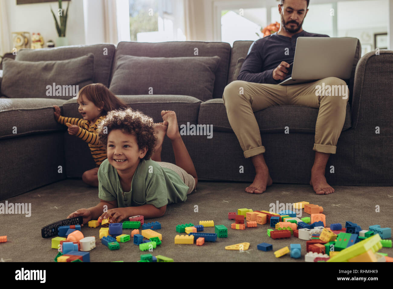 Mann bei der Arbeit am Laptop zu Hause sitzen mit Kinder spielen auf dem Boden. Fröhliche Kinder spielen mit Bausteinen und Fernsehen zu Hause. Stockfoto