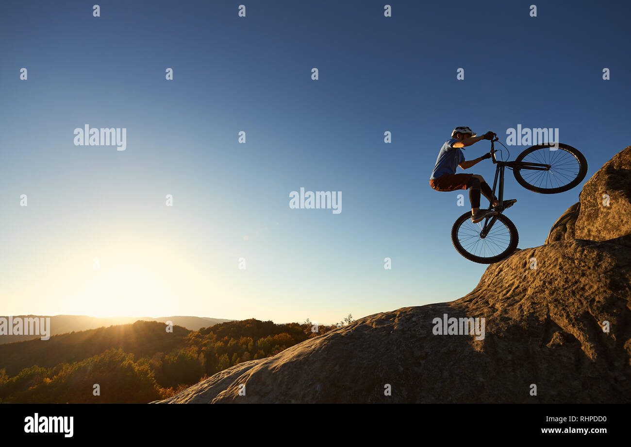 Radfahrer stehen auf der Rückseite Rad Trial Bike. Professionelle Sportler Reiter die akrobatischen Stunts am Rande des großen Felsen auf dem Gipfel des Berges bei Sonnenuntergang. Konzept der Extreme Sport aktiven Lebensstil Stockfoto