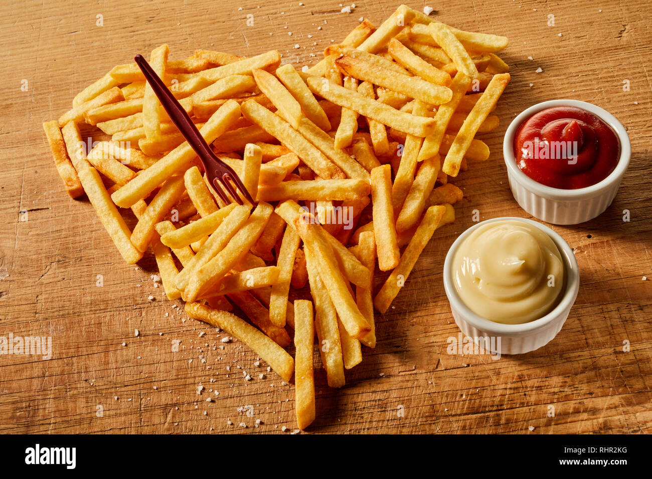 Gesalzen und gewürzt in Herzform servieren von frittierten Kartoffelchips mit Senf und Ketchup auf Holz mit Kunststoff Gabel Stockfoto