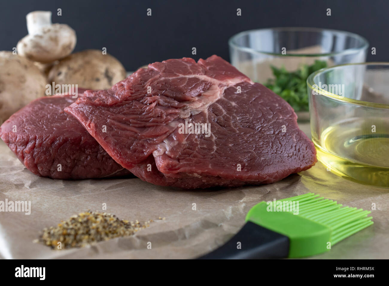 Rohe Steaks prepped für das Kochen. Gewürze im Vordergrund, Pilze im Hintergrund. Schwarzen Hintergrund. 2 Glasschüsseln mit Olivenöl und Petersilie Stockfoto