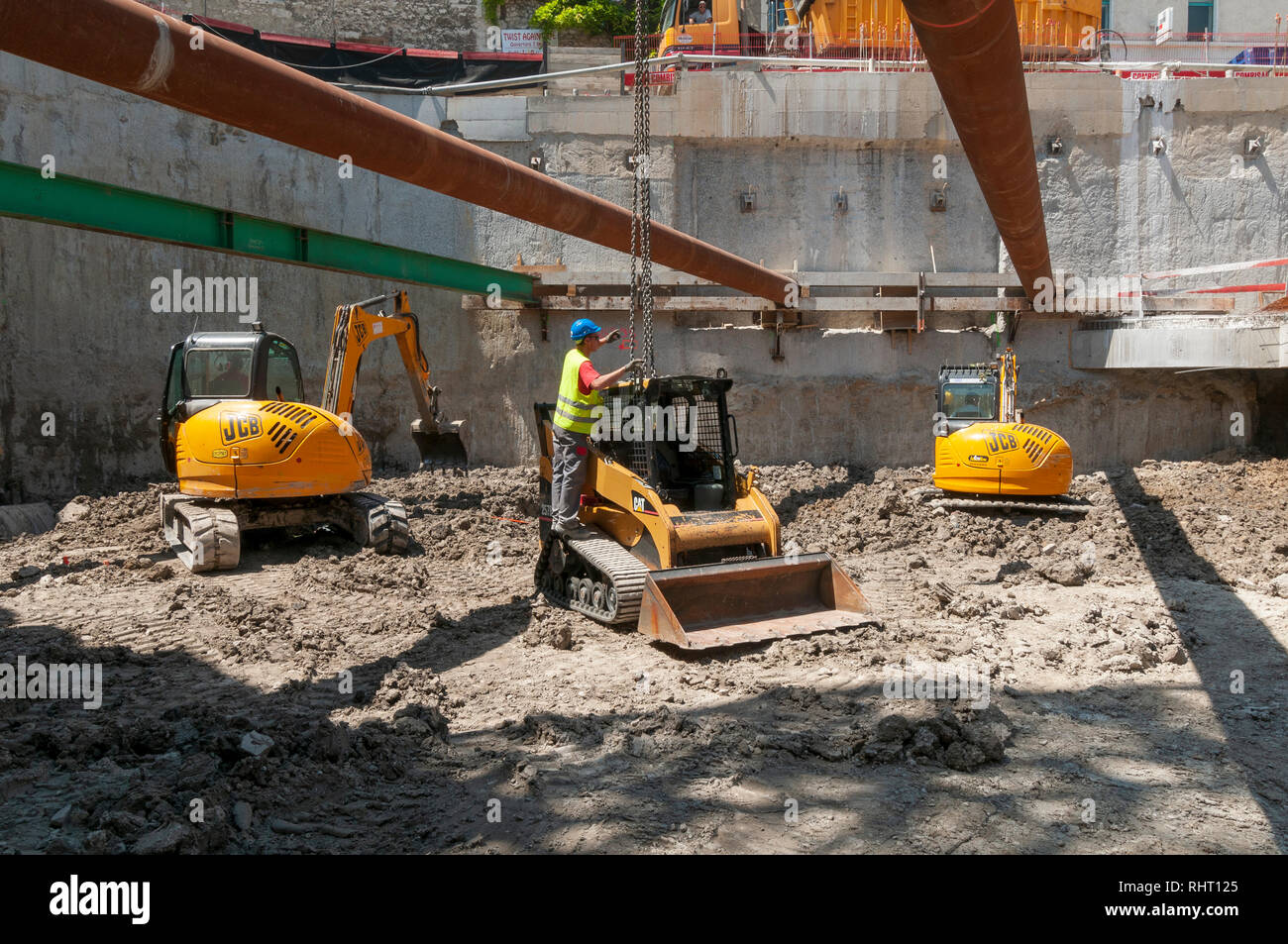 Ein Arbeiter befreit die Ketten von den Kran, dass eine Katze kompakten Kettenlader auf seinen Arbeitsbereich gesenkt hat. Im Hintergrund zwei JCB kompakten Hydraulikbagger. Stockfoto