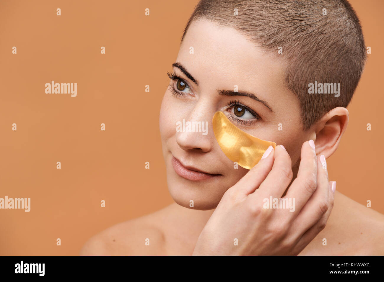 Lächelnd Mitte 30 Frau Anwendung gold Collagen Augenmaske. Foto von attraktiven kaukasische Frau mit Kopf rasiert und gesunde Haut isoliert auf beige backgro Stockfoto
