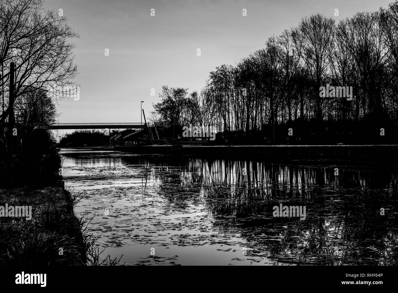 Dramatische und Schwarzweiß Sonnenaufgang über einem schönen frühen Winter Landschaft mit einem gefrorenen Fluss oder Kanal, mit Bäumen gesäumten Riverside und Gras bei Sonnenaufgang Creatin Stockfoto
