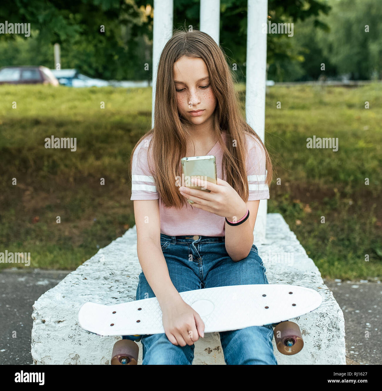 Jugendlich Mädchen 12-13 Jahre alt und sitzt in Händen eines Skate. Im Sommer in der Stadt in lässigen Jeans und pink T-Shirt. Kommunikation im Internet, in die Hände der Stockfoto