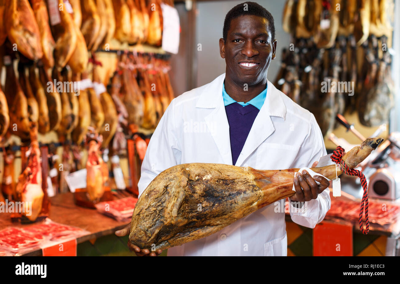 Lächelnd Afrikanische amerikanische Verkäufer in Fleisch shop arbeiten, empfehlen köstliche iberischen Schinken Stockfoto