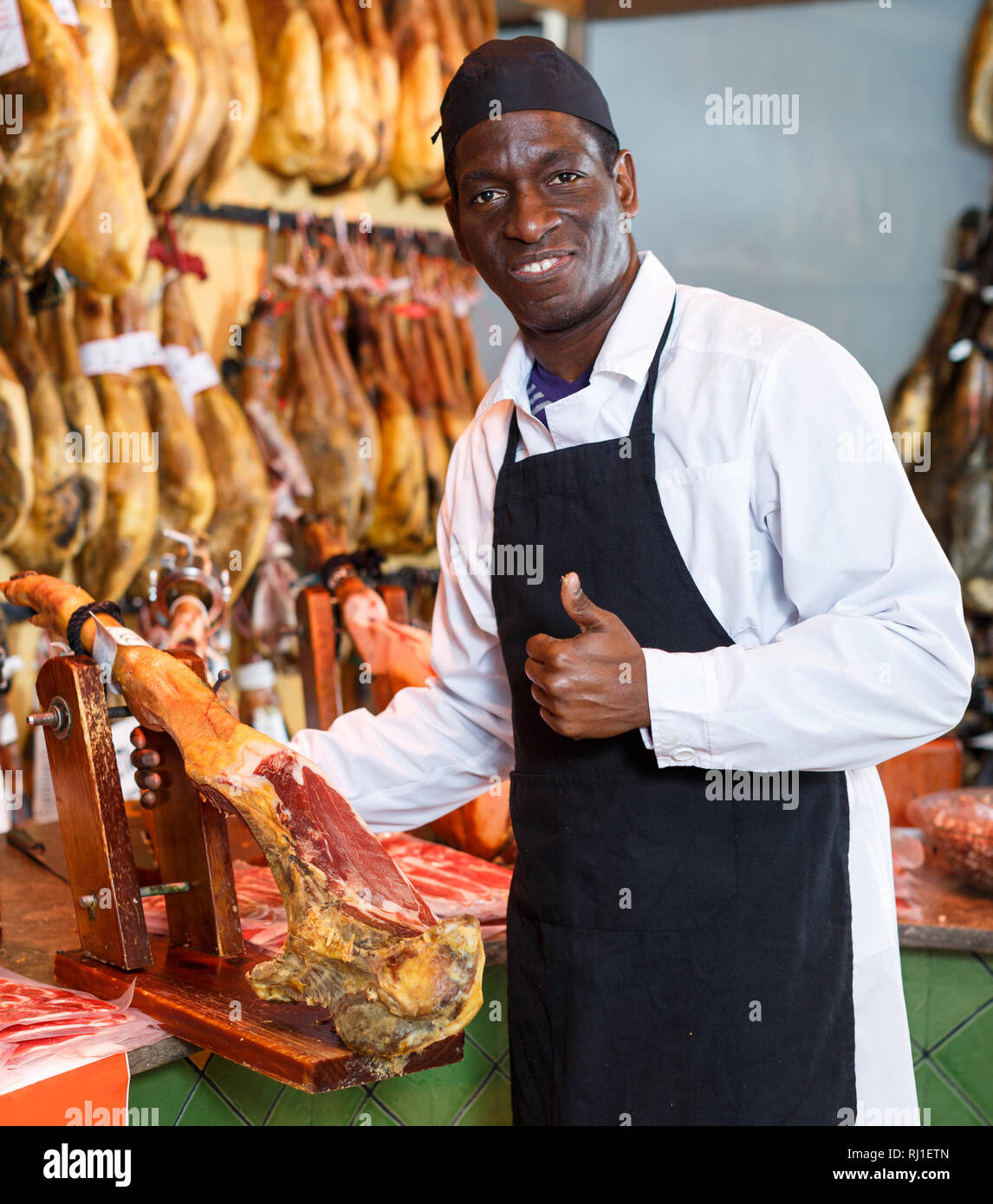 Fröhlich Afrikanische amerikanische Verkäufer zeigen, Daumen hoch und bietet köstliche spanische Jamon in Metzgerei Stockfoto