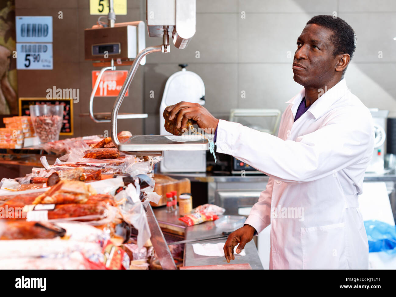 Sicher gerne Fröhliche positive Afrikanische amerikanische Verkäufer wiegen Jamon auf Skala in Metzgerei Stockfoto