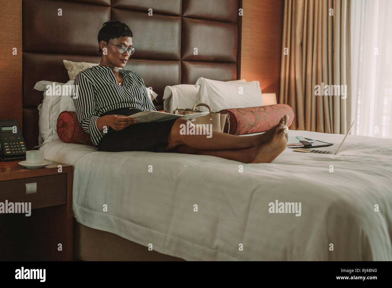 Frau in formelle Kleidung sitzen auf dem Bett mit Zeitung. Geschäftsfrau eine Zeitung lesen in der modernen Hotelzimmer. Stockfoto