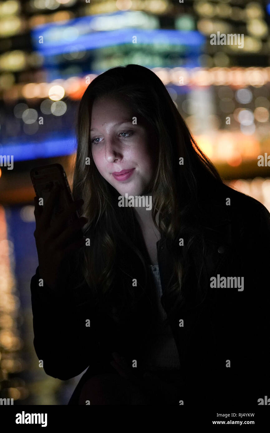 Ein junges Mädchen Gesicht ist von Ihrem Smartphone mit der Skyline der Stadt die Lichter aus in der Ferne geblasen beleuchtet. Stockfoto