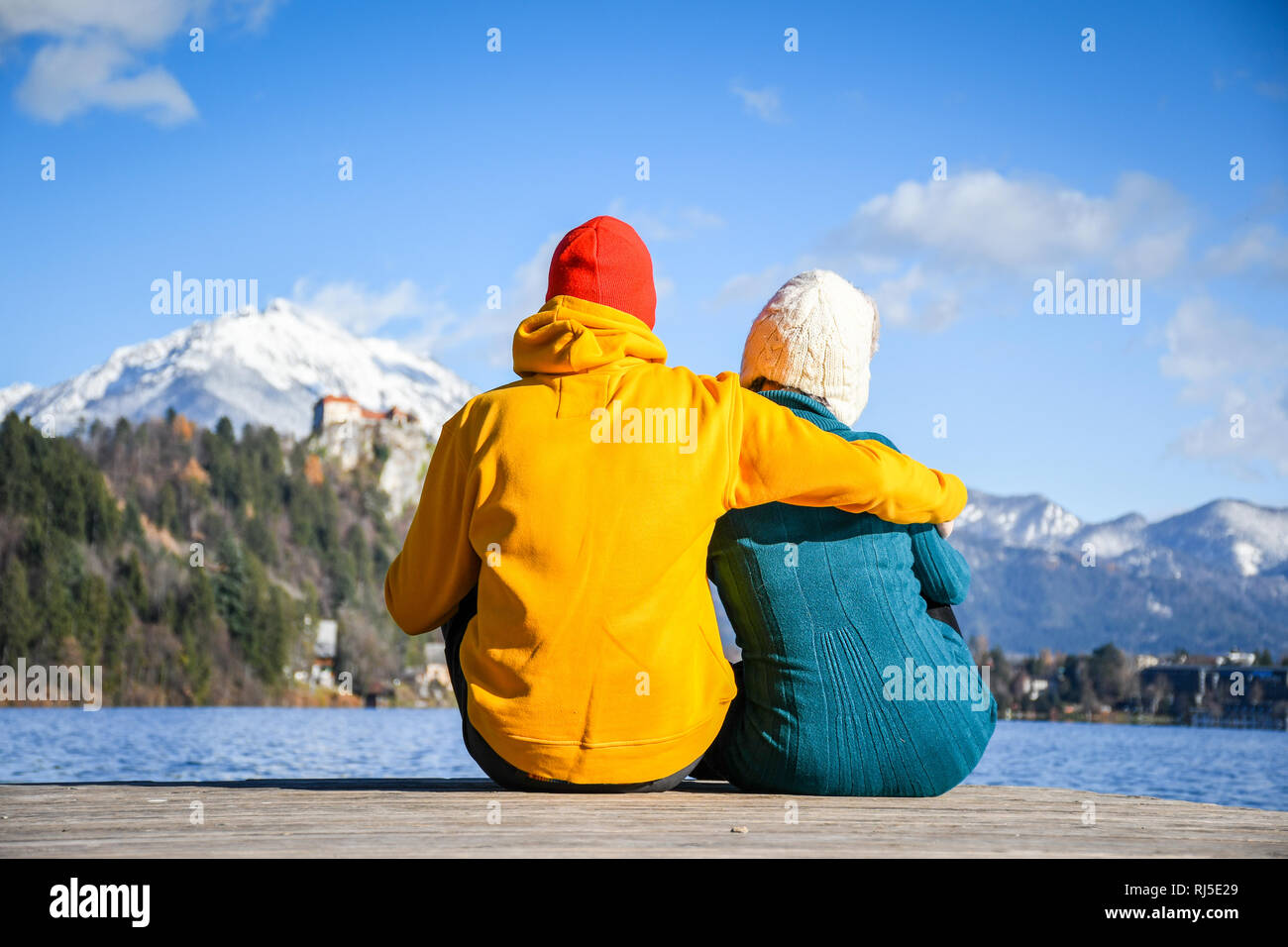 Paar in Liebe umarmen zusammen mit bunten Tüchern sitzen und entspannen auf einer hölzernen Pier auf einem klaren Himmel sonnigen Wintertag Ansicht von der Rückseite, Bled Stockfoto