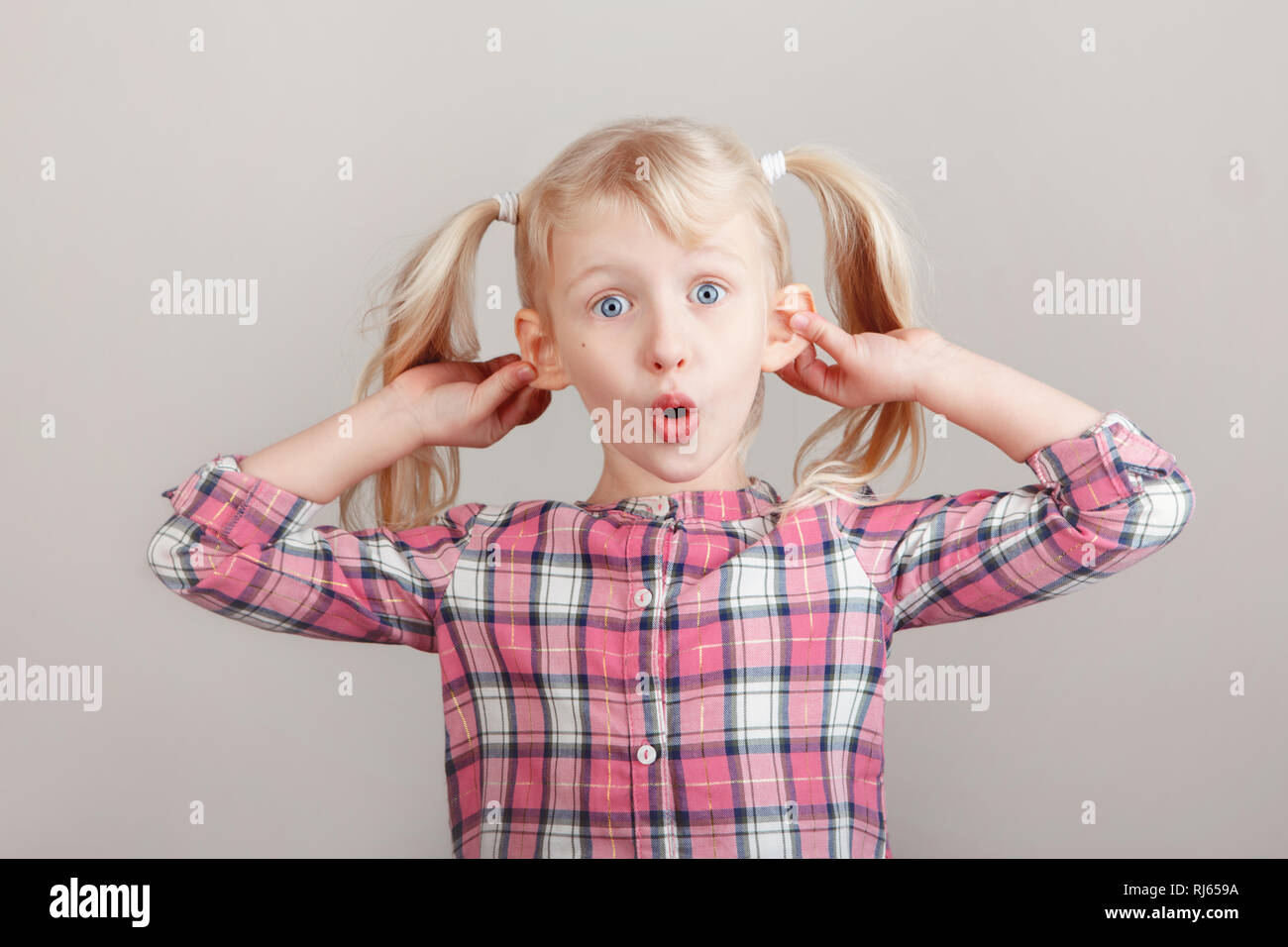 Closeup Portrait von Lustige blonde Kaukasischen Vorschule Mädchen, die Gesichter vor der Kamera. Kind Holding ziehen ziehen Ohren auf hellen Hintergrund. Ki Stockfoto