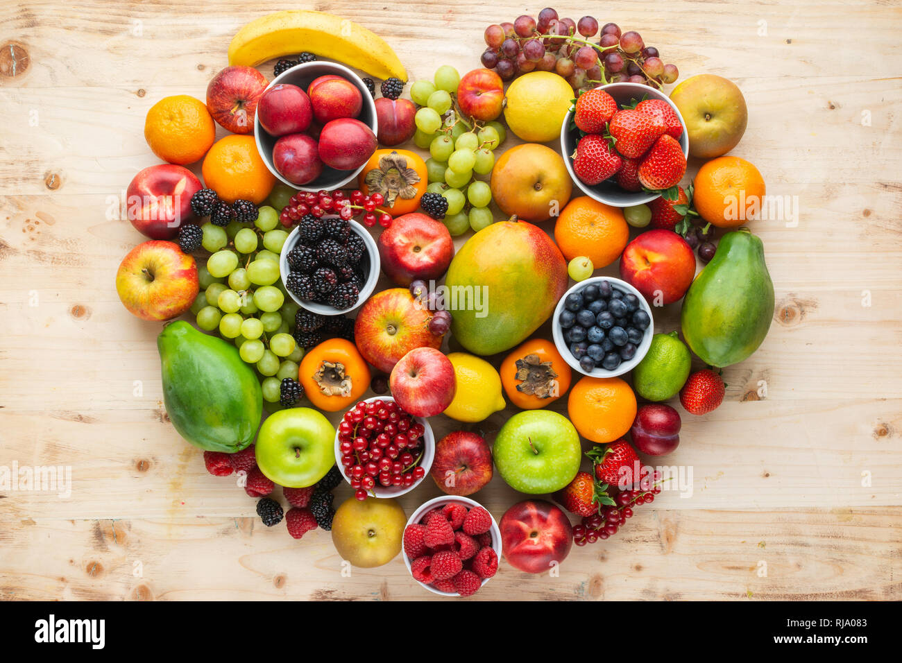 Herz aus gesunden rainbow Früchte, Erdbeeren Himbeeren orangen Pflaumen Äpfel, Kiwis, Weintrauben, Heidelbeeren mango Kaki Ananas auf hellem Holz. Stockfoto