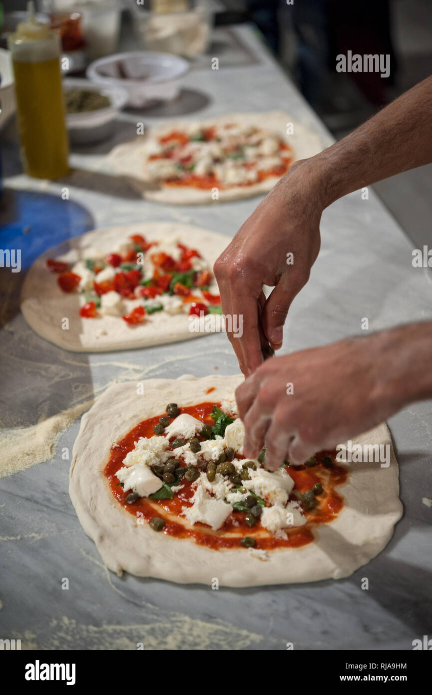 Vorbereitung Pizza Margherita auf einem Ablageflächen aus Marmor. Pizzaiolo setzt Stück Mozzarella, Kapern und Basilikum über eine rohe Pizza Teig. Stockfoto