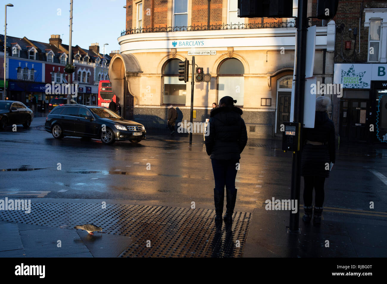 Auf der Straße beobachten, wie Leute über ihr tägliches Leben mit Sonnenschein und Reflexionen auf dem Bürgersteig an einem regnerischen Tag gehen in Leytonstone im Osten von London, Vereinigtes Königreich. Leytonstone ist ein Gebiet im Osten von London, und ein Teil der Londoner Stadtteil Waltham Forest. Stockfoto