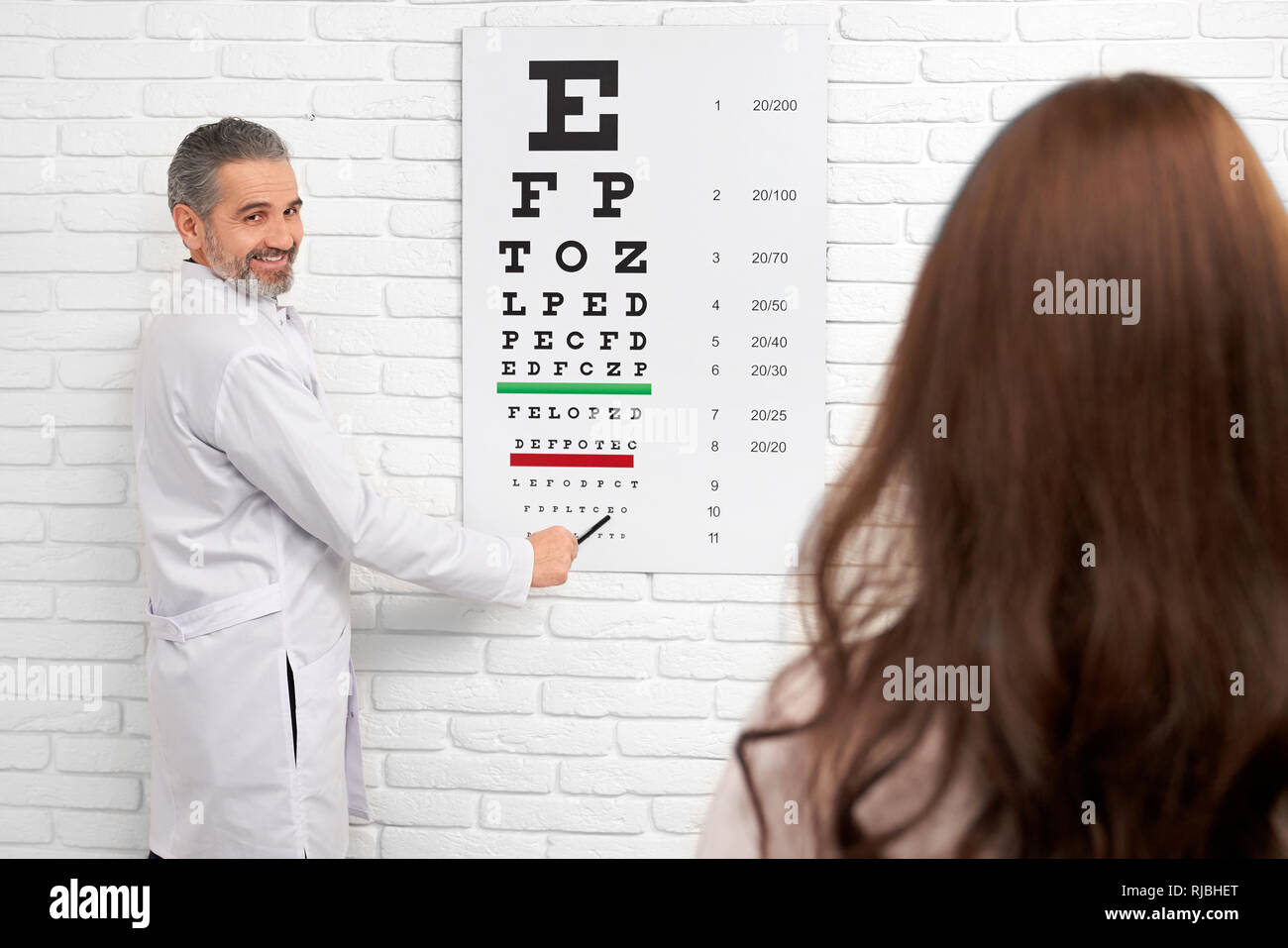 Professionelle Augenarzt am Auge Diagramm zeigend, Überprüfung des Sehvermögens in der Klinik. Fröhlich lächelnd, Augenarzt konsultieren bei Patienten suchen. Augenarzt tragen in Weiß medizinische Fell. Stockfoto