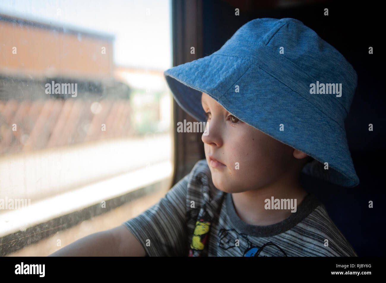 Ein kleiner Junge mit Engelsgesicht, der mit einem blauen Hut verziert ist, fesselt die Reise mit Wunderaugen und blickt aus dem Zugfenster. Stockfoto