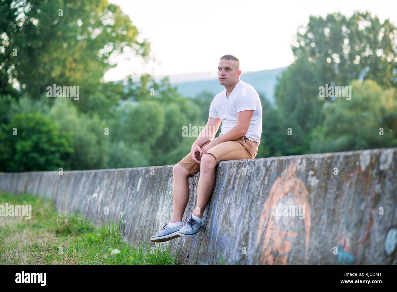 Junge einsame Mann sitzt auf einer Steinmauer im sonnigen Sommer Natur. Stockfoto