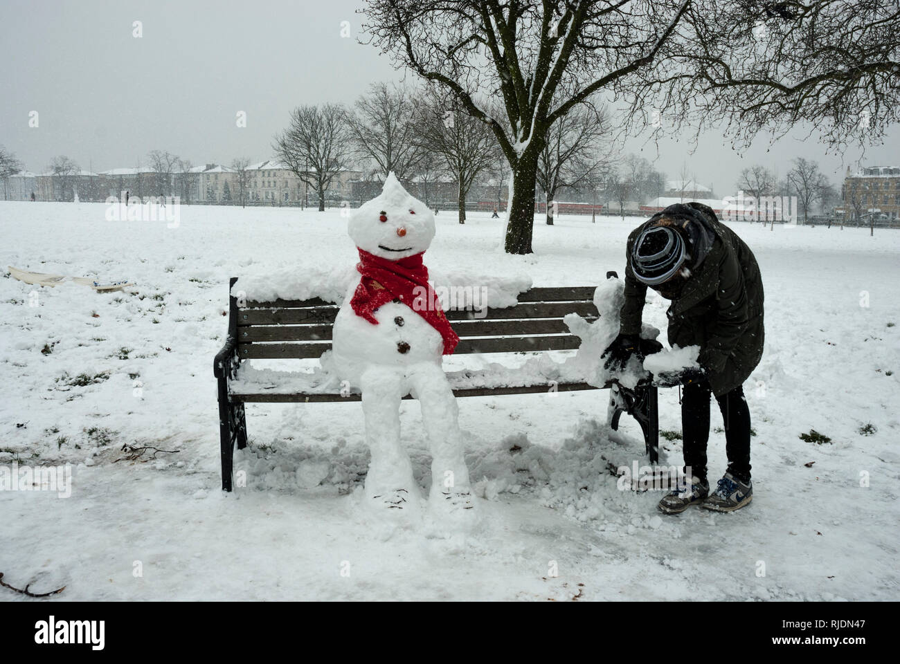 Haringey Finsbury Park, London, UK. Einen fröhlichen Schneemann entspannend auf einer Bank mit einer jungen Person, die einem Schnee baby Daneben und Schnee herum. Stockfoto
