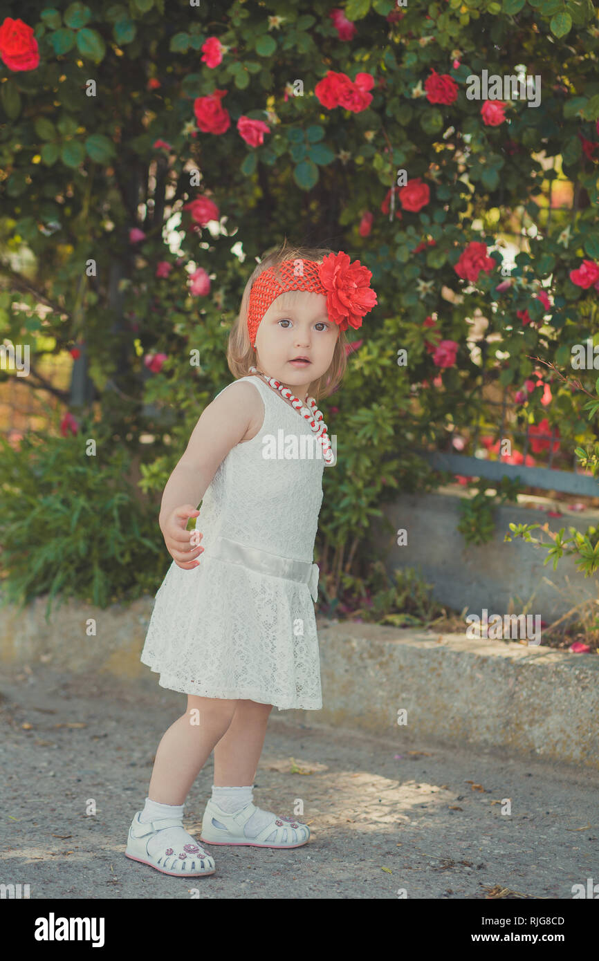 Retro stilvoll gekleideten blonden jungen Baby Mädchen im Central Park Garten posing tragen Französische couturer weißes Kleid rotes Halstuch und Rosenkranz. Stockfoto