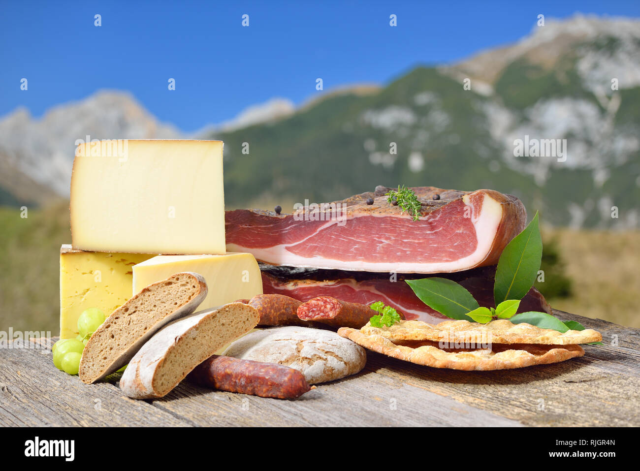 Südtiroler Spezialitäten wie Speck, wurst und Käse liegen auf einem rustikalen Tisch vor der Berge der Alpen. Stockfoto