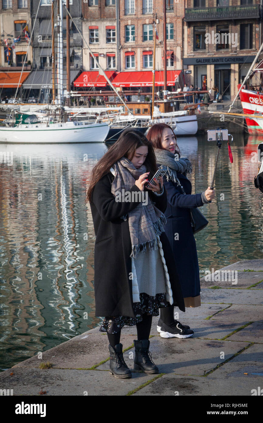 Ein paar der jungen Frauen aus einer japanischen touristische Gruppe bzw. schicken Sie eine SMS, und nehmen Sie eine selfie durch den berühmten Hafen von Honfleur, Normandie, Frankreich. Stockfoto