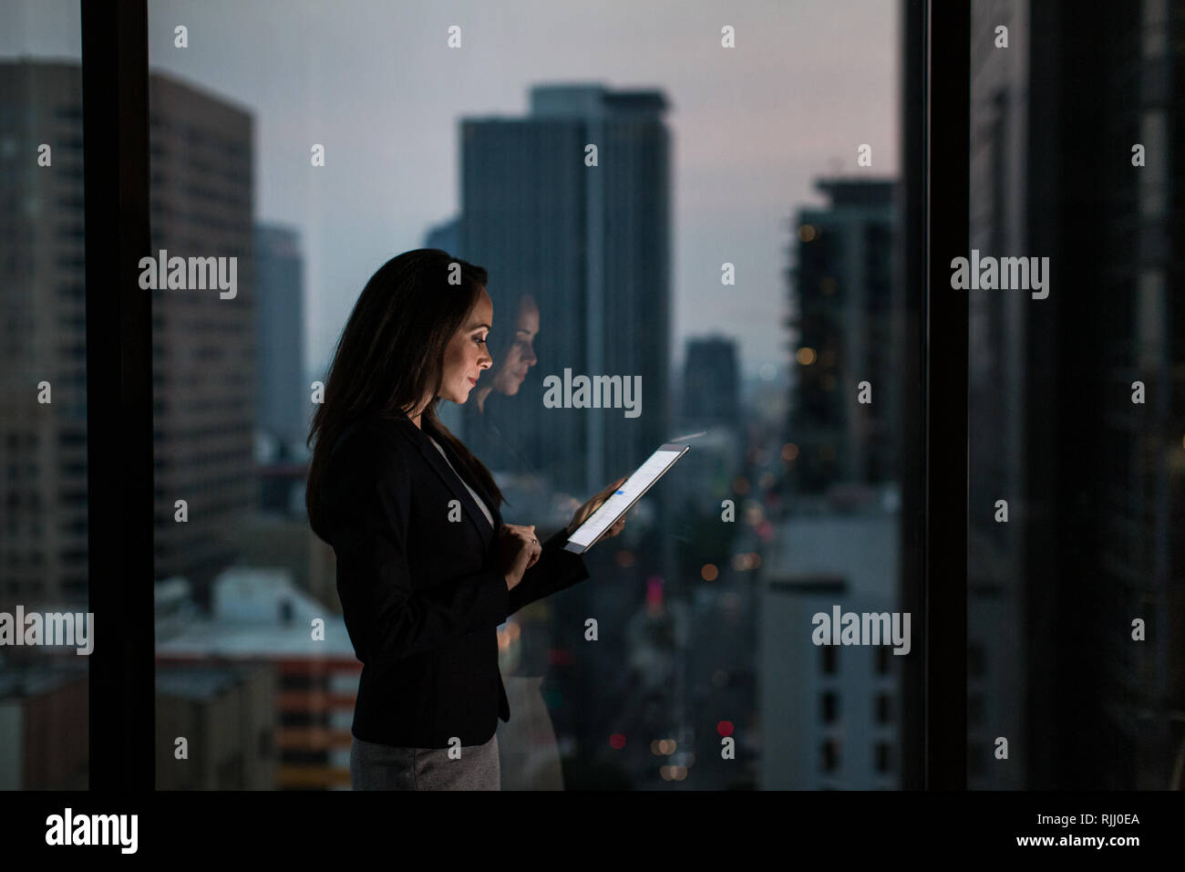 Geschäftsfrau, die spät mit digitalen Tablet mit Sicht auf die City Skyline im Hintergrund Stockfoto
