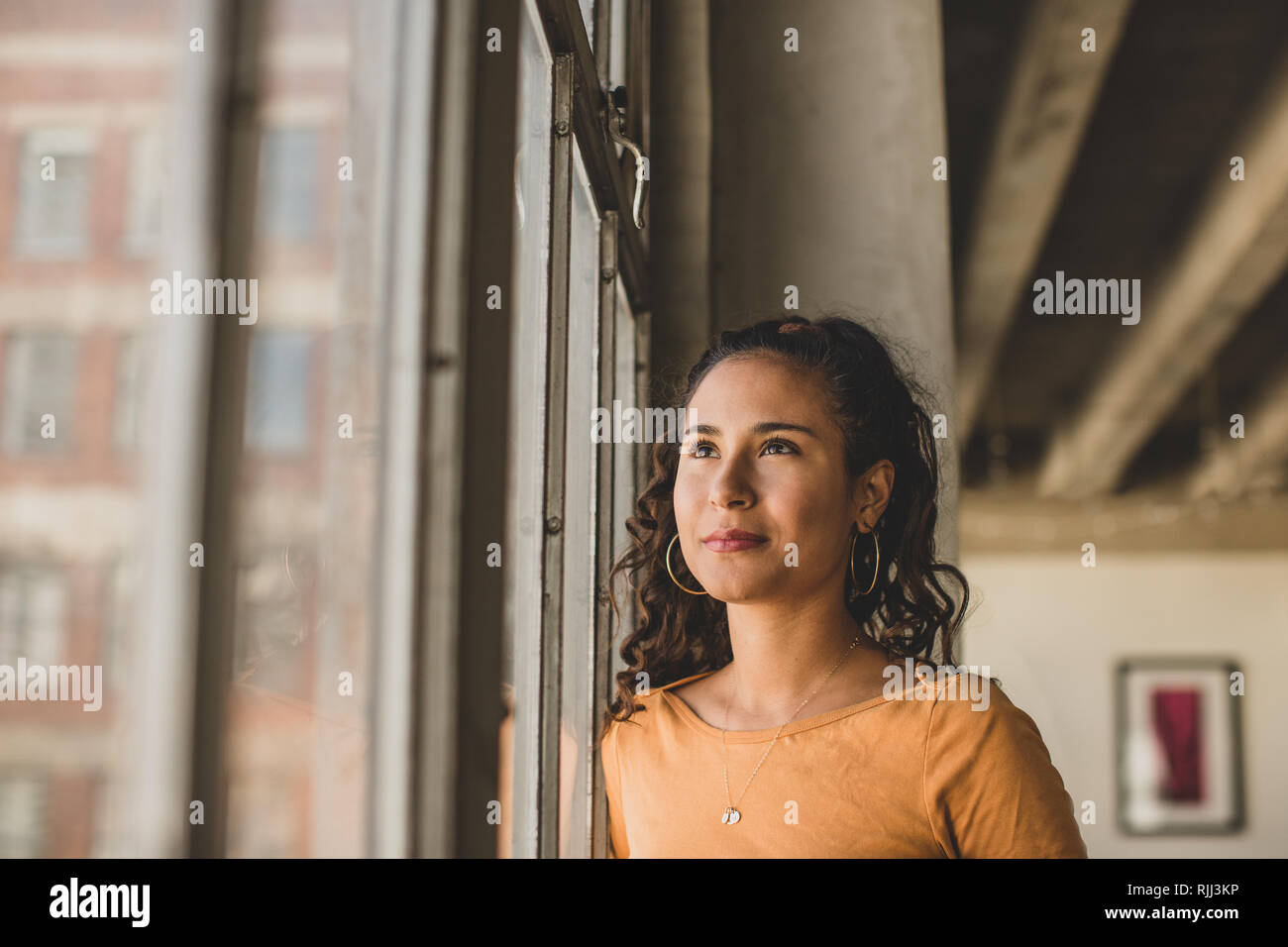 Junge Hispanic weiblichen Blick aus Fenster in loft apartment Denken Stockfoto